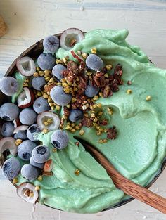 a bowl filled with blueberries and green smoothie next to a wooden spoon on top of a white table