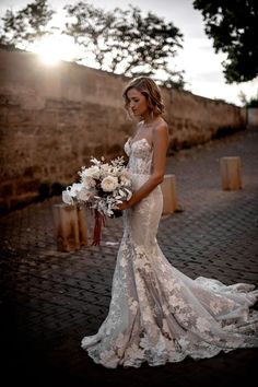 a woman in a wedding dress is holding a bouquet and posing for the camera on a cobblestone street