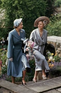 two women in dresses and hats are walking down the steps to an outdoor garden area