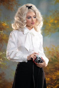 A blond pinup model posing against a yellow and blue painted background while wearing the Angela Blouse in white and black trousers with an old camera on her hand. Fall Puff Sleeve Blouse With Bow, Fall Puff Sleeve Blouse With Bow Detail, Elegant Fall Blouse With Bow Tie, White Fall Blouse With Bow, Fall White Blouse With Tie Sleeves, White Tie Sleeve Blouse For Fall, White Tie Sleeves Blouse For Fall, White Blouse With Tie Sleeves For Fall, Classic Fitted Blouse With Bow