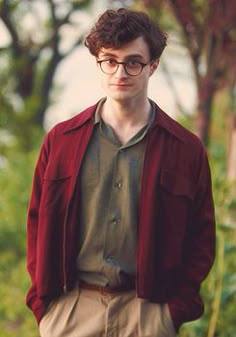 a young man wearing glasses and a red jacket standing in front of some trees with his hands in his pockets