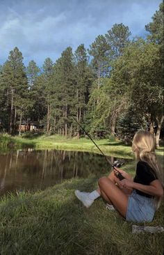 a woman sitting on the grass with a fishing rod in her hand while she sits next to a pond