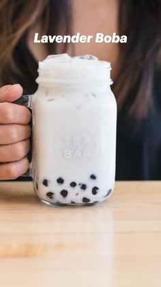 a woman holding a mason jar filled with liquid and coffee beans on top of a wooden table