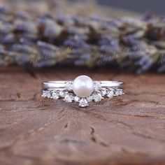 a white pearl and diamond ring sitting on top of a piece of wood with lavender flowers in the background