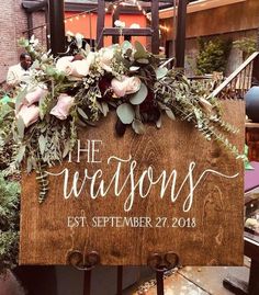 a wooden sign with flowers and greenery hanging from it's sides in front of a brick building