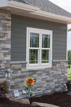 a flower in front of a gray house with white windows and brick siding on the side