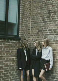 three women standing next to each other near a brick building with windows on the side