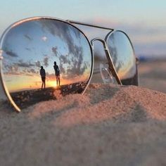 two people standing in the sand with their reflection in them's sunglasses, as the sun sets behind them