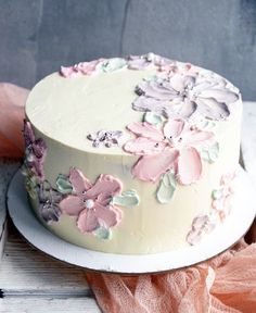 a white cake with pink and purple flowers on it sitting on a table next to a piece of cloth