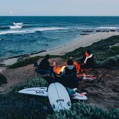 people sitting around a campfire on the beach with surfboards in front of them