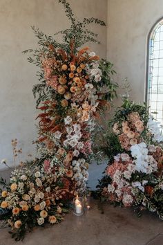 an arrangement of flowers and greenery on the floor next to a candle in front of a window