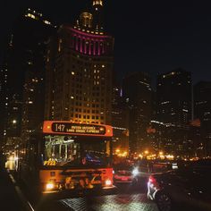 the city bus is driving down the street at night with its lights on and buildings lit up in the background