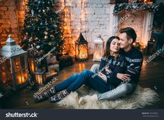 a young couple sitting on the floor in front of a christmas tree with lights and presents