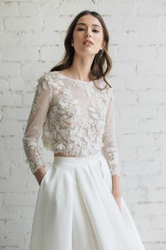 a woman wearing a white wedding dress with sheer sleeves and flowers on the top, standing in front of a brick wall