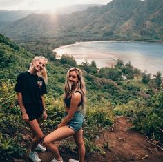 two young women standing on top of a hill next to a body of water and mountains