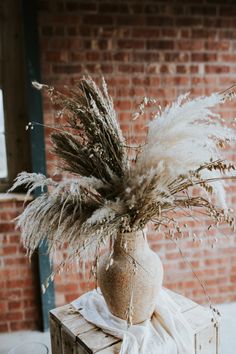 a vase filled with dry grass sitting on top of a wooden box next to a brick wall