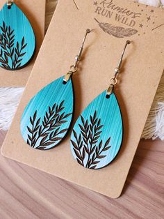 two pairs of earrings with blue and black leaves painted on the outside of them, sitting on top of a piece of cardboard