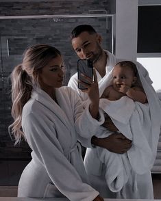 a man and woman taking a selfie in the bathroom with their baby wrapped in white robes