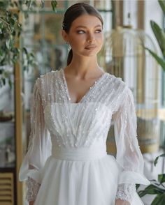 a woman wearing a white dress with long sleeves and beading on the shoulders is standing in front of a plant