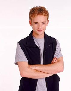 a young man with his arms crossed standing in front of a white background wearing a black vest