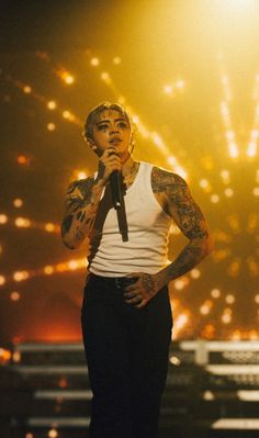 tattooed man in white tank top and black pants on stage holding microphone with fireworks behind him