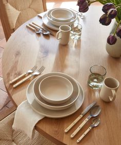 a wooden table topped with white plates and silverware next to a vase filled with purple flowers