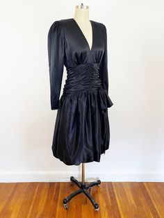 "Dance the night away in this flirty black satin bubble dress. Made by Hildegarde Sausik for Lalique Designs circa 1980s. Black polyester satin. Broad shoulder pad shoulders, tapered long sleeves, deep v-neck, fitted bodice and nipped at the waist with gathered detail that accentuates the waist. The bubble skirt has tule inside, hidden, for a full look. The back is stunning open with button at the neck, fully lined and back nylon zipper. In beautiful vintage condition, no flaws to note, ready to wear.  Measurements Estimated modern women's size Small Shoulder to Shoulder 15\" Shoulder to Sleeve Cuff 24\" Bust 40\" Natural Waist 28\" Waist Meets Skirt 34\" Hips (skirt lining) 38\" Length Shoulder to Waist 23\" Length Waist to Skirt Hem 23\" All items are pre-owned and show signs of wear or 80s Party Dress, 80s Goth, Satin Noir, 80s Party, Goth Dress, Bubble Dress, Bubble Skirt, 80s Dress, Full Look