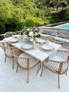 an outdoor dining table with white plates and place settings next to a swimming pool surrounded by greenery