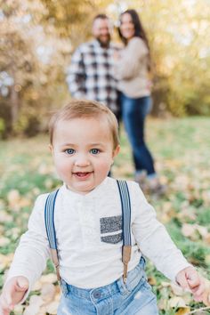 a baby boy in overalls and suspenders smiles at the camera while his parents stand behind him