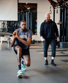 two men in a gym doing squats with one man looking at the camera while another watches