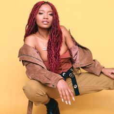 a woman with red dreadlocks sitting on a stool wearing a brown jacket and khaki pants