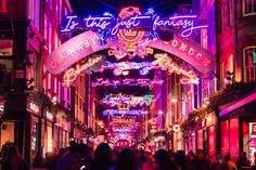 people are walking down the street at night with neon signs above them and buildings in the background