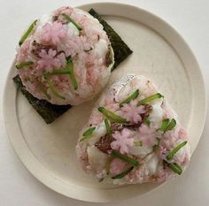 two pieces of sushi on a white plate with green onions and pink flower garnishes