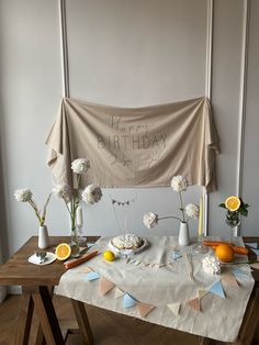 a table with flowers and fruit on it in front of a happy birthday banner hanging from the wall