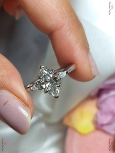 a woman's hand holding a diamond ring on top of her finger with flowers in the background