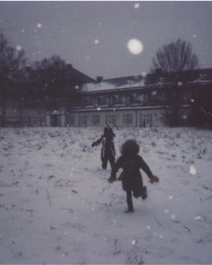 two children are playing in the snow near a house and trees, with one child running towards the camera