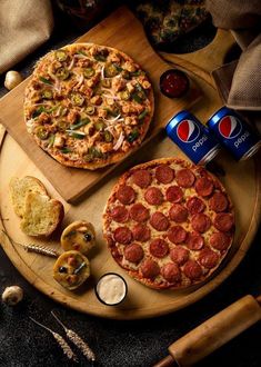 two pepperoni pizzas on a wooden platter next to cans of soda and bread