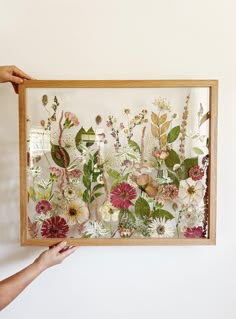 a person holding up a framed artwork with flowers and leaves on the wall behind it