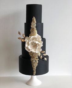 a three tiered black cake with white flowers on the top and gold leaf decorations