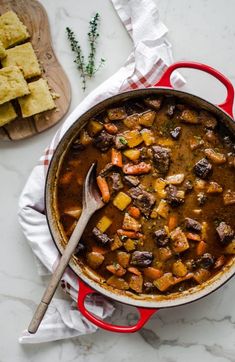 a red pot filled with stew next to a wooden cutting board and serving utensils