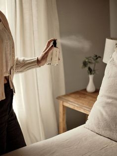 a woman holding a bottle in her hand while standing next to a bed with white sheets