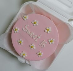 a pink frosted cake with white flowers on it sitting in a plastic container next to a spoon