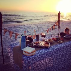 two people sitting at a table on the beach with food and drinks in front of them