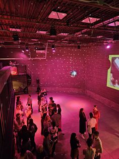 a group of people standing around in a room with pink lights on the ceiling and a projector screen behind them