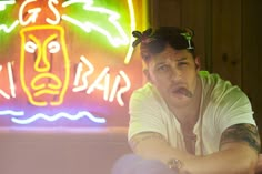 a man leaning on a table in front of a neon sign