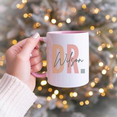 a person holding a pink and white coffee mug with the word dr on it in front of a christmas tree