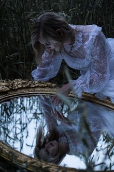 a woman in white dress leaning over a mirror with grass and plants around her reflection