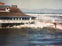 a large wave crashes into the ocean near a restaurant and pier on a cloudy day