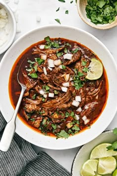 a white bowl filled with beef and onions next to bowls of cilantro, limes and rice