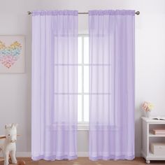 a white teddy bear sitting in front of a window with purple sheer curtains on it